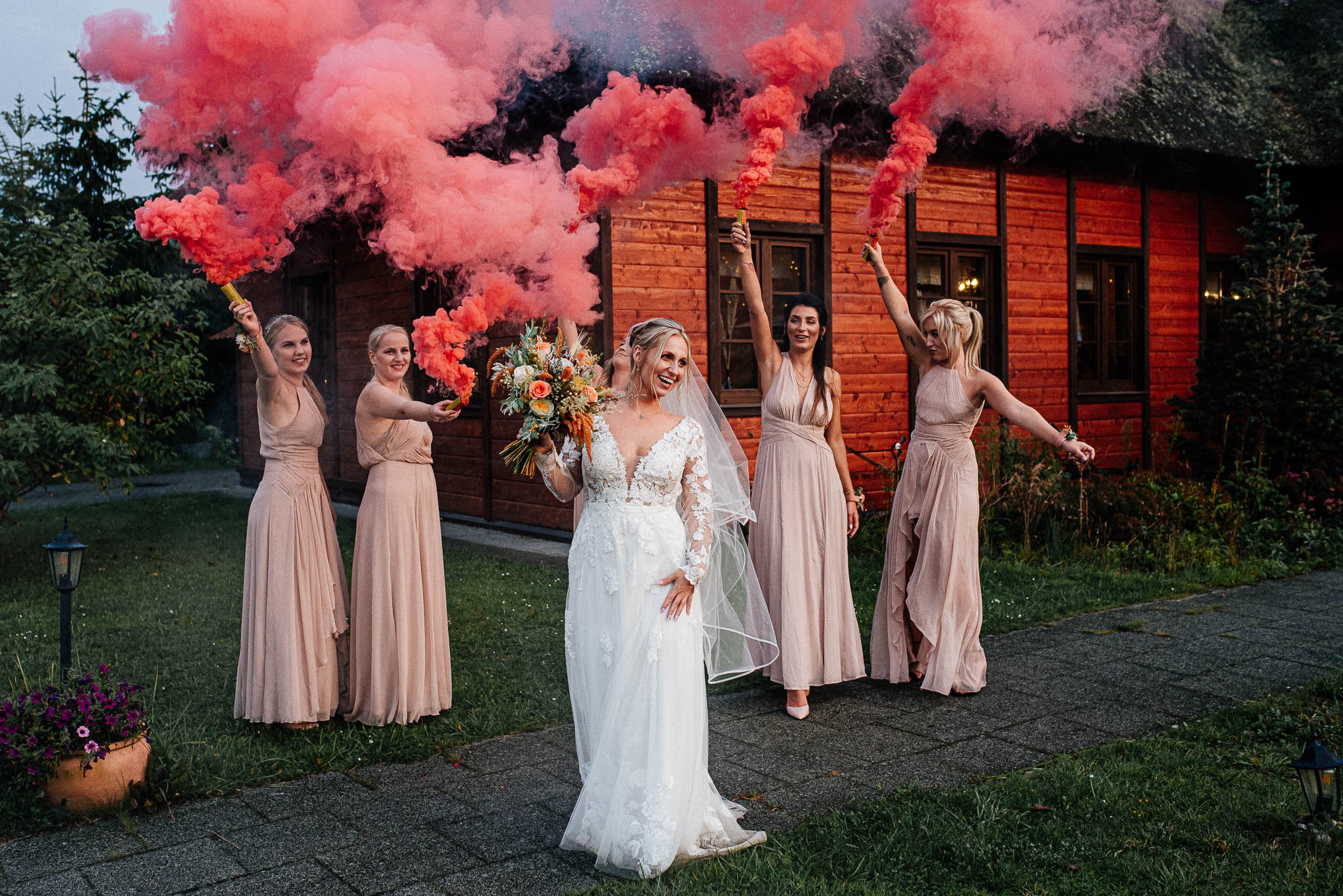 Na zdjęciu widzimy panią młodą w towarzystwie swoich druhen podczas sesji ślubnej z użyciem świec dymnych. W tle widać rustykalną chatę, która tworzy piękne tło dla fotografii.